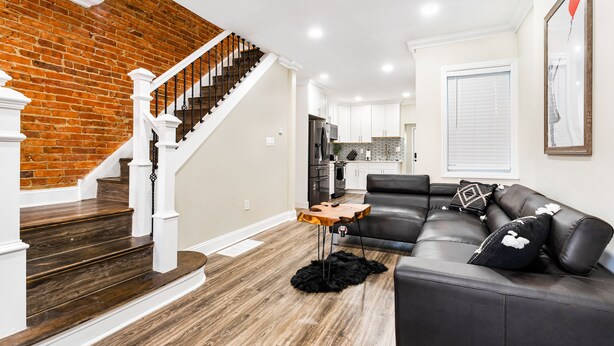A modern living room with a dark wood staircase, a leather sofa, and a kitchen in the background.