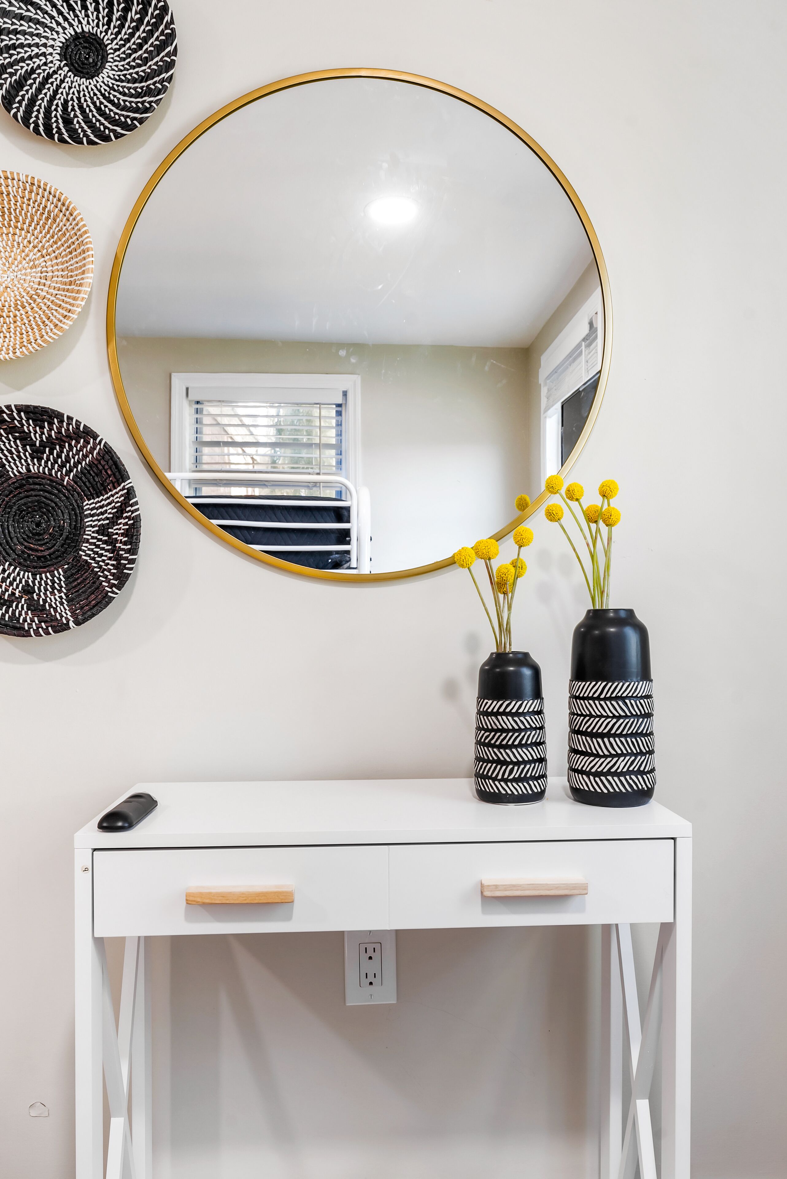 A white desk with two black vases with white patterns, a round mirror with a gold frame, and a wall-mounted decorative plate.