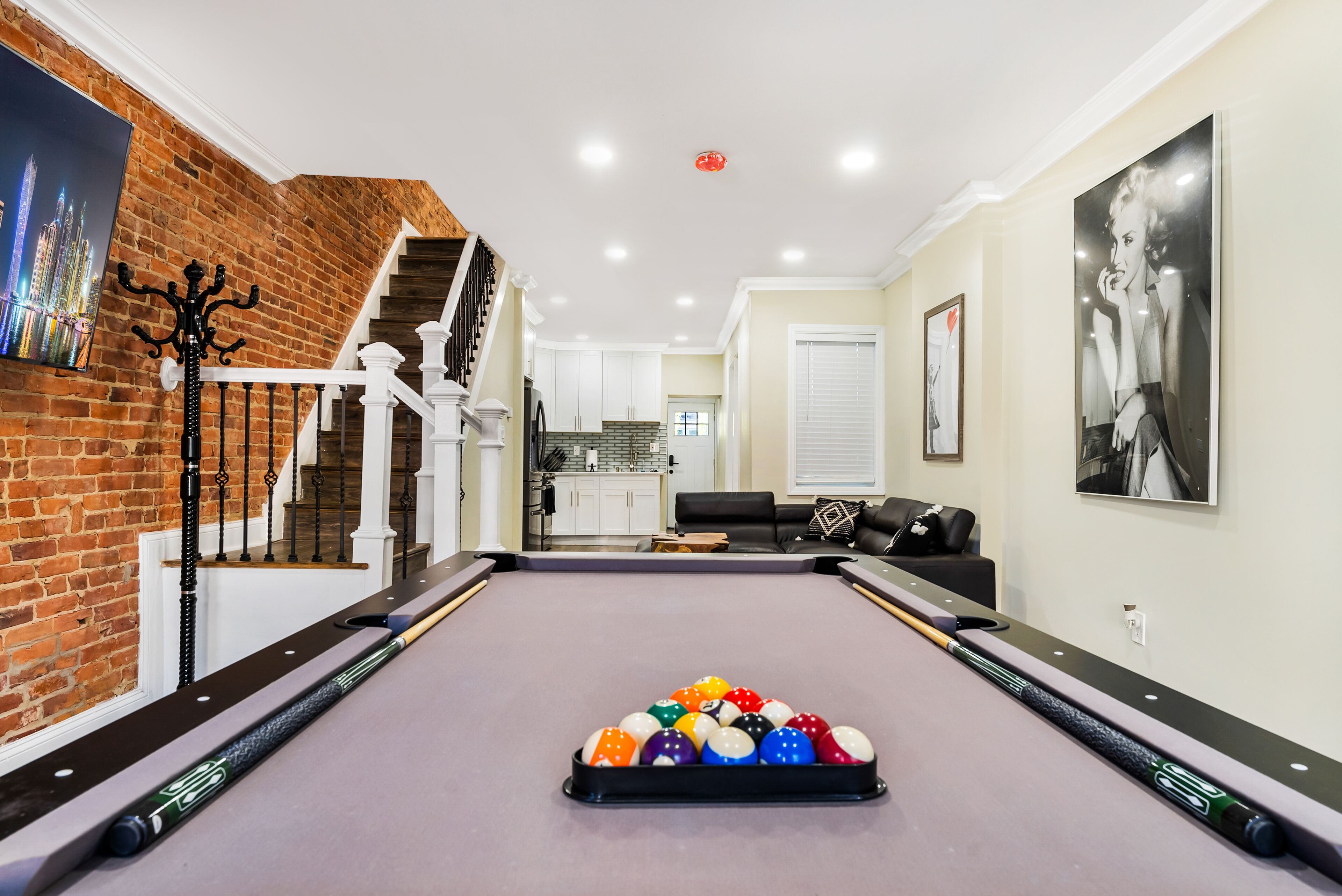 A pool table with balls, a staircase, a kitchen, and a living area with a couch and artwork.