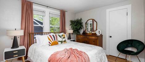 Un dormitorio con cama, mesitas de noche, cómoda, silla, espejo y ventana con cortinas.