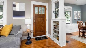 A well-lit living room with a grey sofa, a wooden door, a small table with a lamp, and a dining area with a wooden table and chairs. - Walk to Shops & Dining, Great Location! - 3br (Perrysburg)