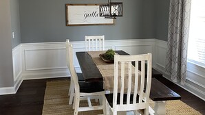 Un comedor con una mesa de madera oscura, sillas blancas, un tapete tejido y una lámpara colgante.
