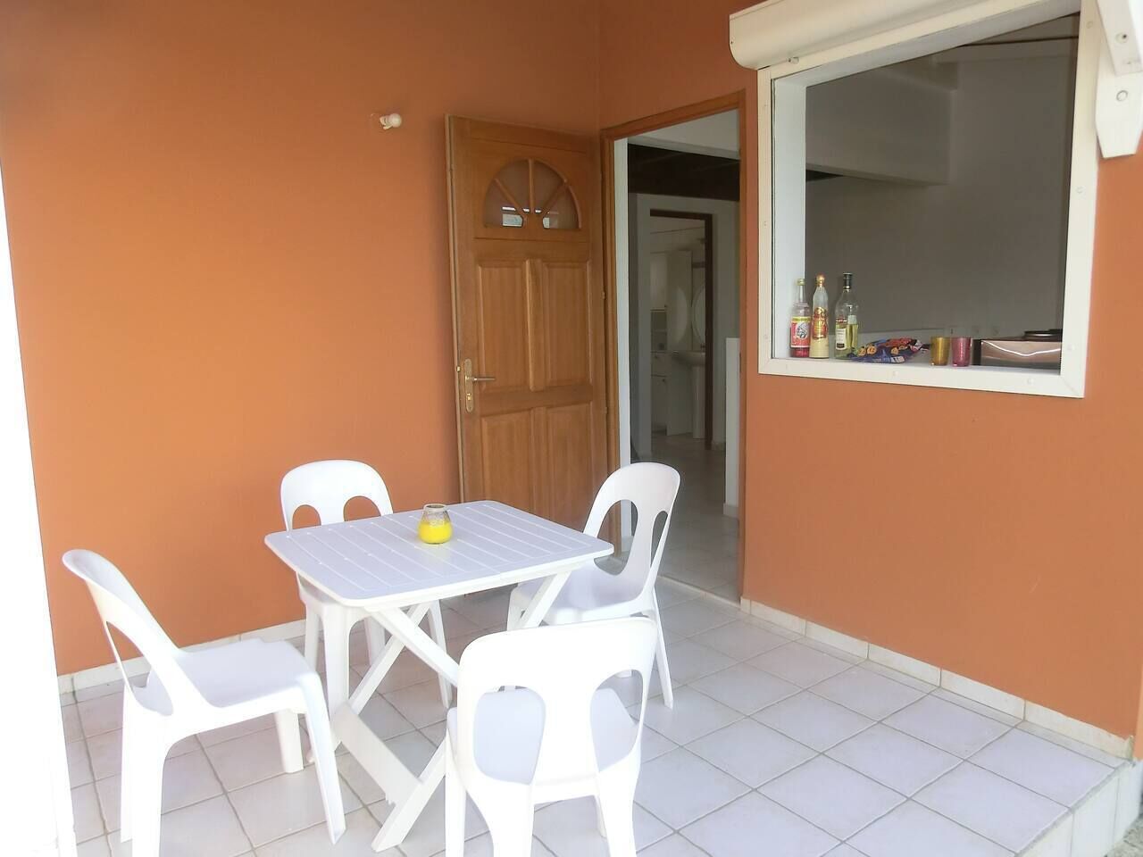 Un petit pati amb una taula blanca i quatre caires blancs, una porta de fusta i una finestra amb ampolles a la finestra.