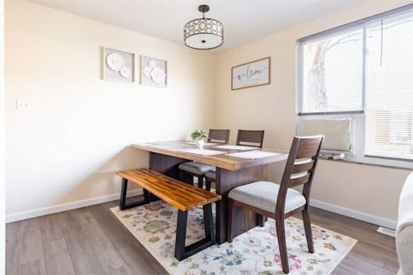 A dining area with a wooden table, chairs, a bench, a chandelier, and a window with blinds.