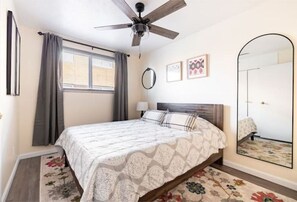 A bedroom with a bed, a ceiling fan, a window with curtains, a mirror, and a floral rug.