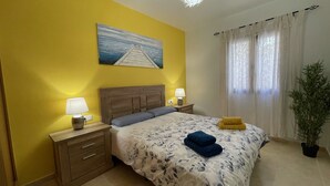 Un dormitorio con cabecera de madera, una cama con ropa de cama floral, dos lámparas de noche, un cuadro de un muelle y una planta en maceta.