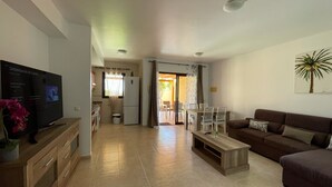 Una sala moderna con televisor de pantalla plana, una mesa de centro de madera, un sofá marrón y una zona de comedor con sillas y una mesa.