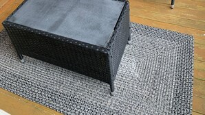 Una habitación con dos sillas de mimbre negro y cojines blancos, un otomano tejido en negro y un suelo de madera.