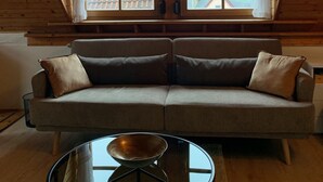 Una habitación acogedora con techo inclinado, un ventanal con cortina de encaje, un sofá beige con cojines y una mesa de centro de vidrio con un cuenco.