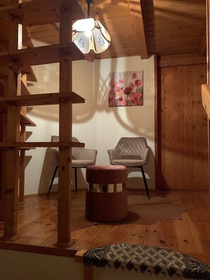 A wooden interior with a ceiling light, two chairs, a round ottoman, and a patterned rug.