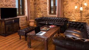 Un salon chaleureux avec un canapé en cuir, une table basse en bois sur laquelle se trouve un ordinateur portable, un téléviseur posé sur un meuble en bois et un mur en pierre.