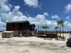 A wooden building with a deck and a small shed, located on a sandy beach with palm trees and a clear sky.