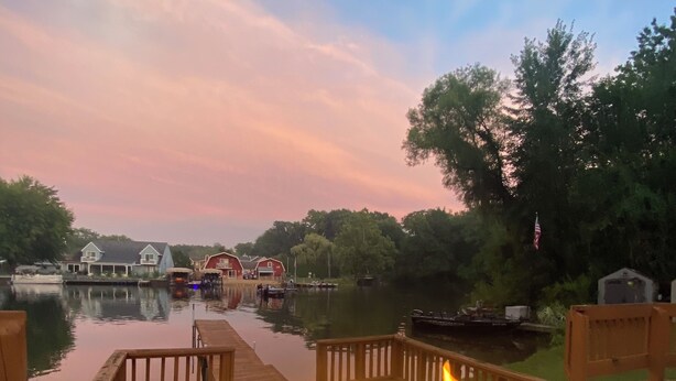 A deck with a fire feature and two glasses of water, overlooking a calm body of water with boats and a dock in the background. - Newly remodeled LAKE HOUSE with HOT TUB, Game Room, Large DECK on PAW PAW LAKE (Coloma)