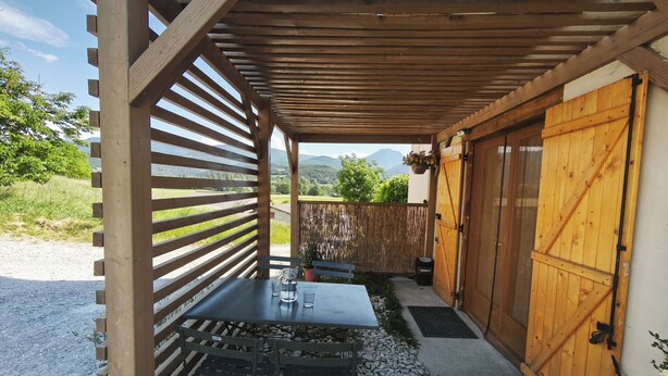 Un espace extérieur abrité, doté d’une table et de bancs, d’une pergola en bois, d’une porte en bois et offrant une vue sur le paysage environnant.