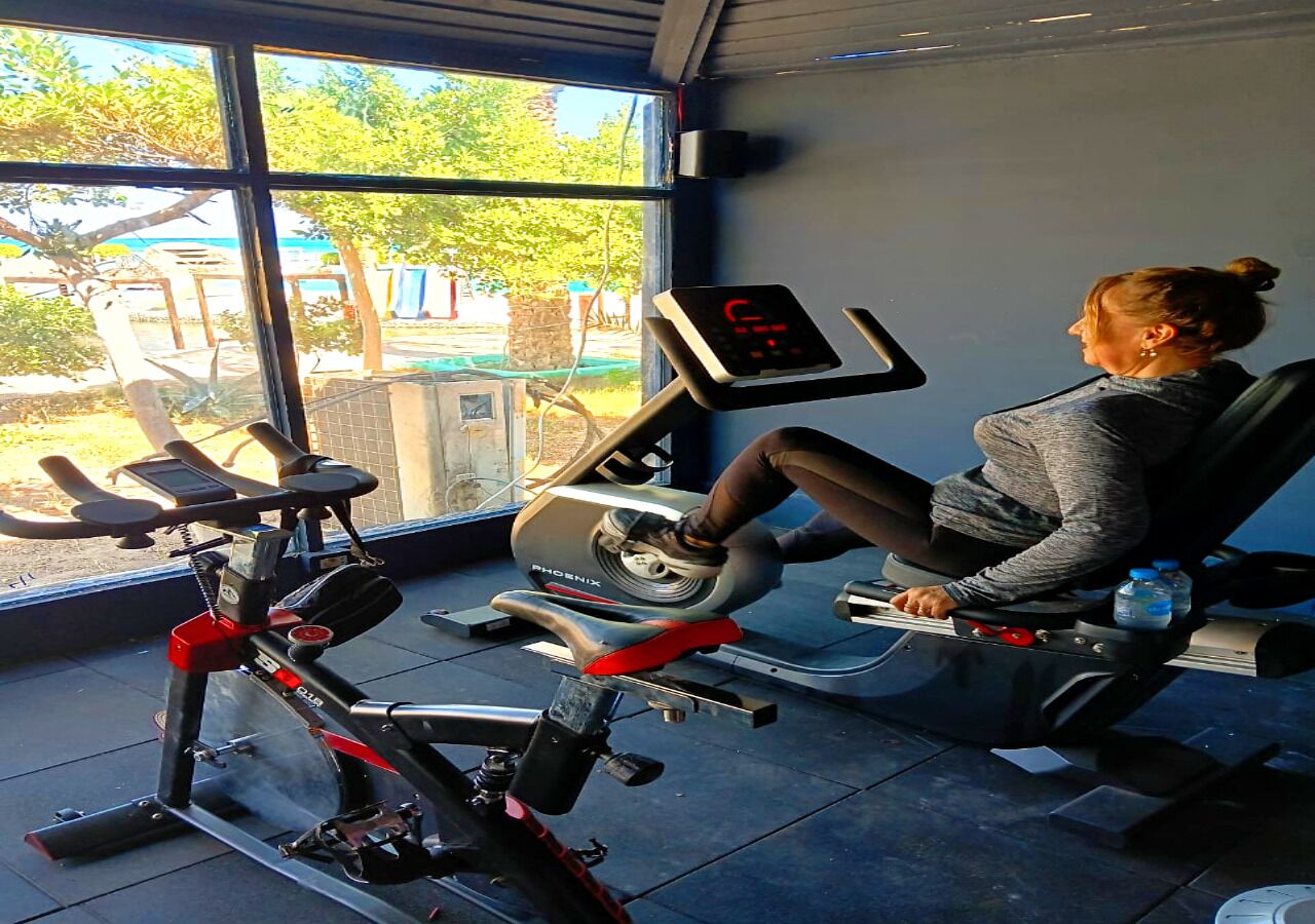 A woman using an exercise bike in a gym with a view of trees and a building outside.