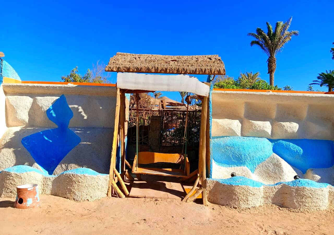 A small, rustic wooden structure with a thatched roof, possibly a seating area or a small shelter, set against a wall with blue and white painted sections.