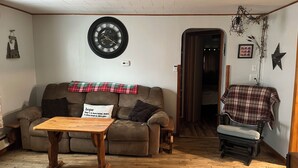 A cozy living room with a wooden floor, a brown sofa with a plaid blanket, a wooden coffee table, and a wall clock. - Cozy Nothwoods home next to ATV/Snowmobile trails (Laona)