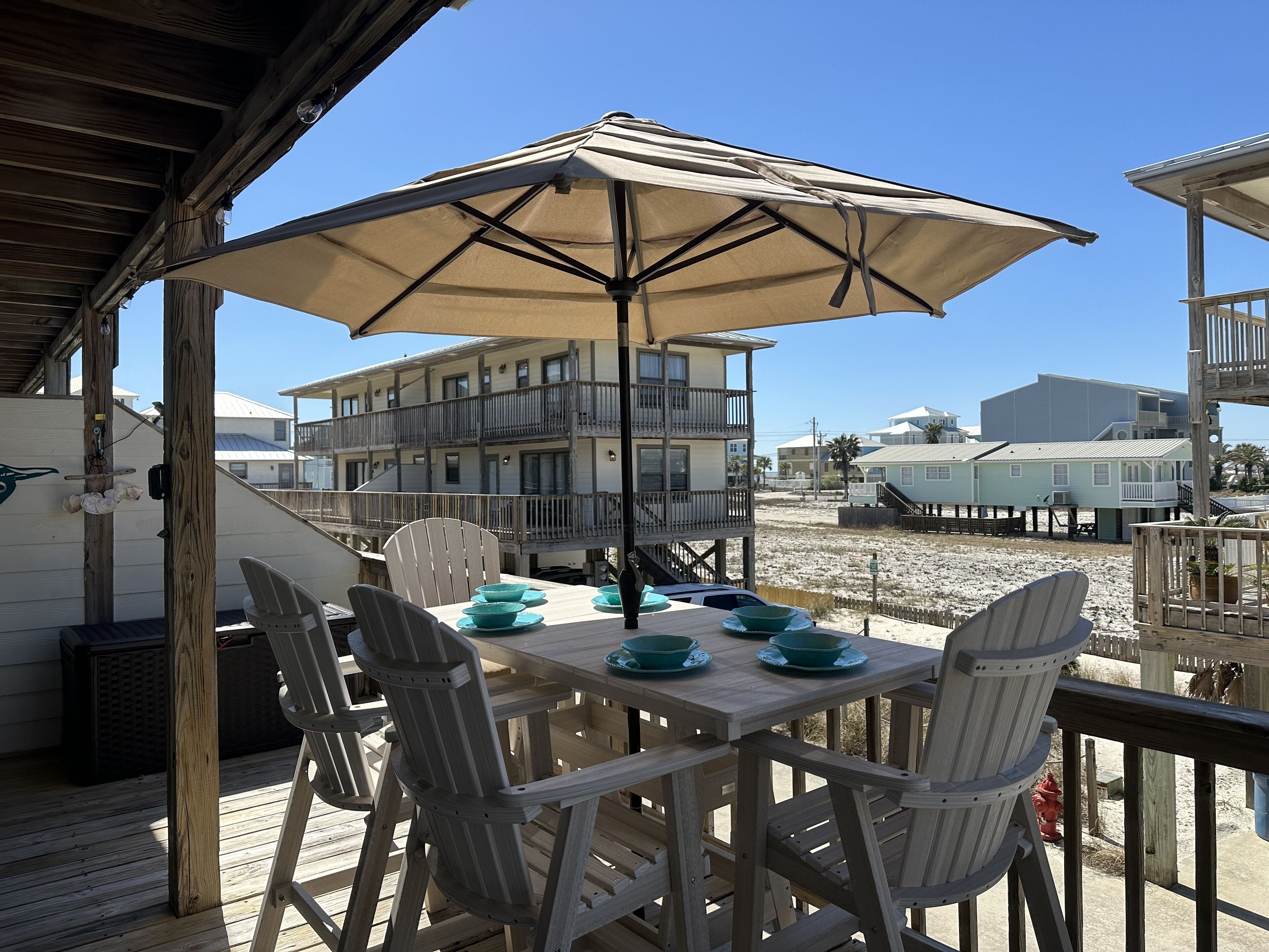 Een houten terras met een tafel, stoelen en een parasol, met op de achtergrond gebouwen en strand.