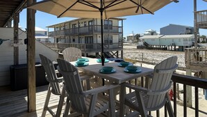 Eine Holzterrasse mit Tisch und Stühlen, einem Sonnenschirm und Blick auf Gebäude und Strand im Hintergrund.