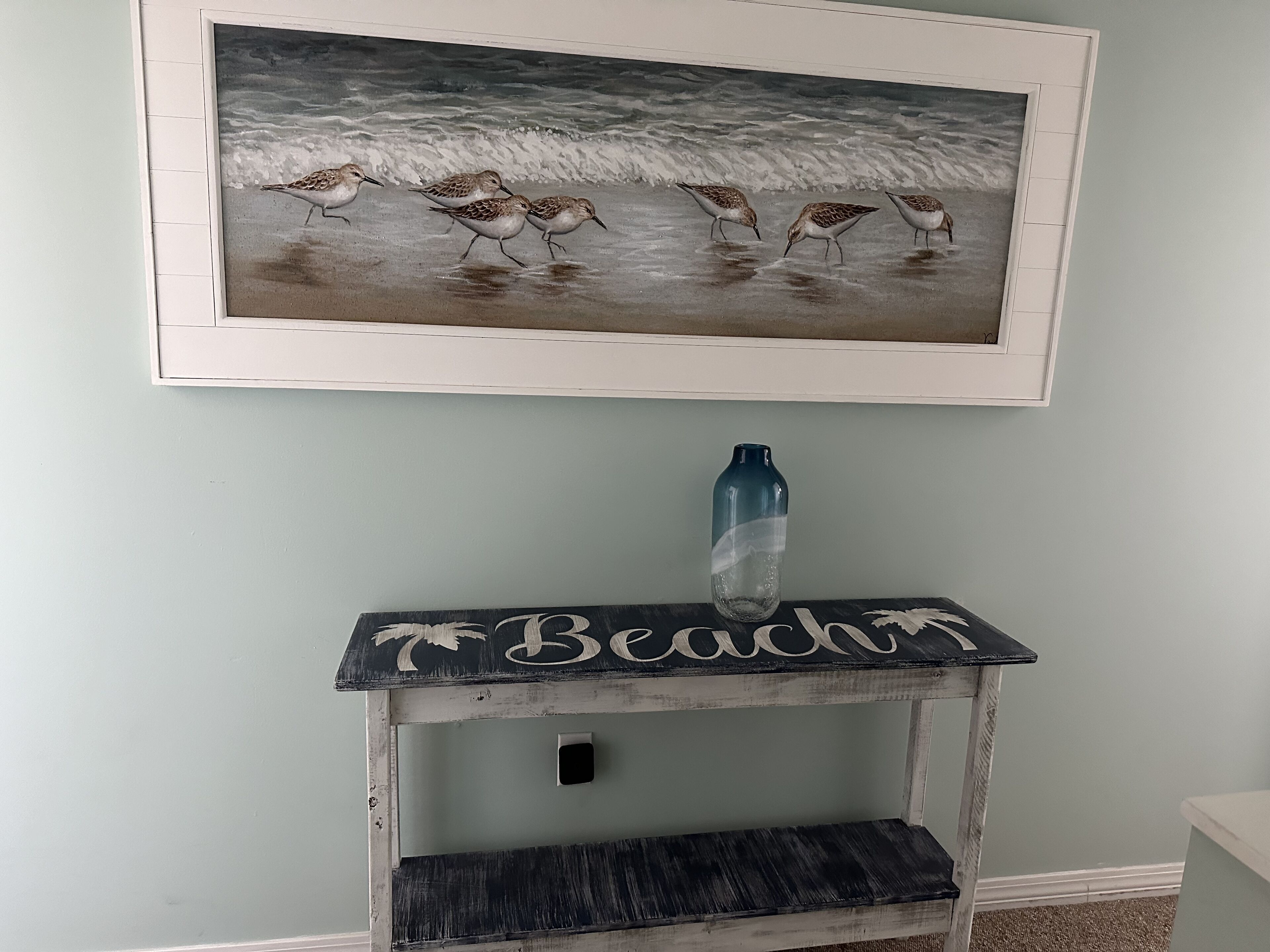 A wall-mounted picture of birds on a beach, a shelf with the word "Beadlin" on it, and a blue bottle.