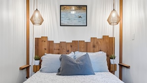 A bed with a patterned bedspread, wooden headboard, two bedside tables with potted plants, and a framed picture above the bed. - Steps from the Beach! ~ Near Wild Pacific Trail! ⎮ Orca (Ucluelet)