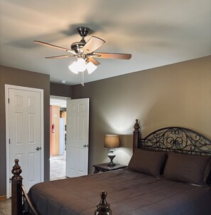 A bedroom with a bed, a nightstand, a lamp, a ceiling fan, and a door leading to another room. - Whispering Oaks Cabin 13 (Oden)