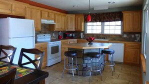 Una cocina con armarios de madera, un refrigerador blanco, una estufa, un microondas y una lámpara colgante roja. Hay una mesa de comedor con sillas y una zona de taburetes de bar.