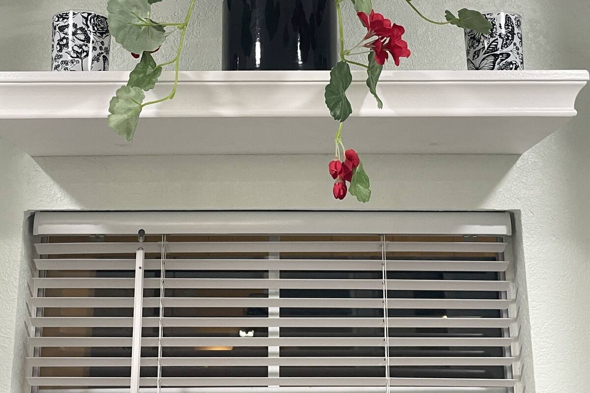A potted plant with red flowers on a white shelf above a window with white blinds.