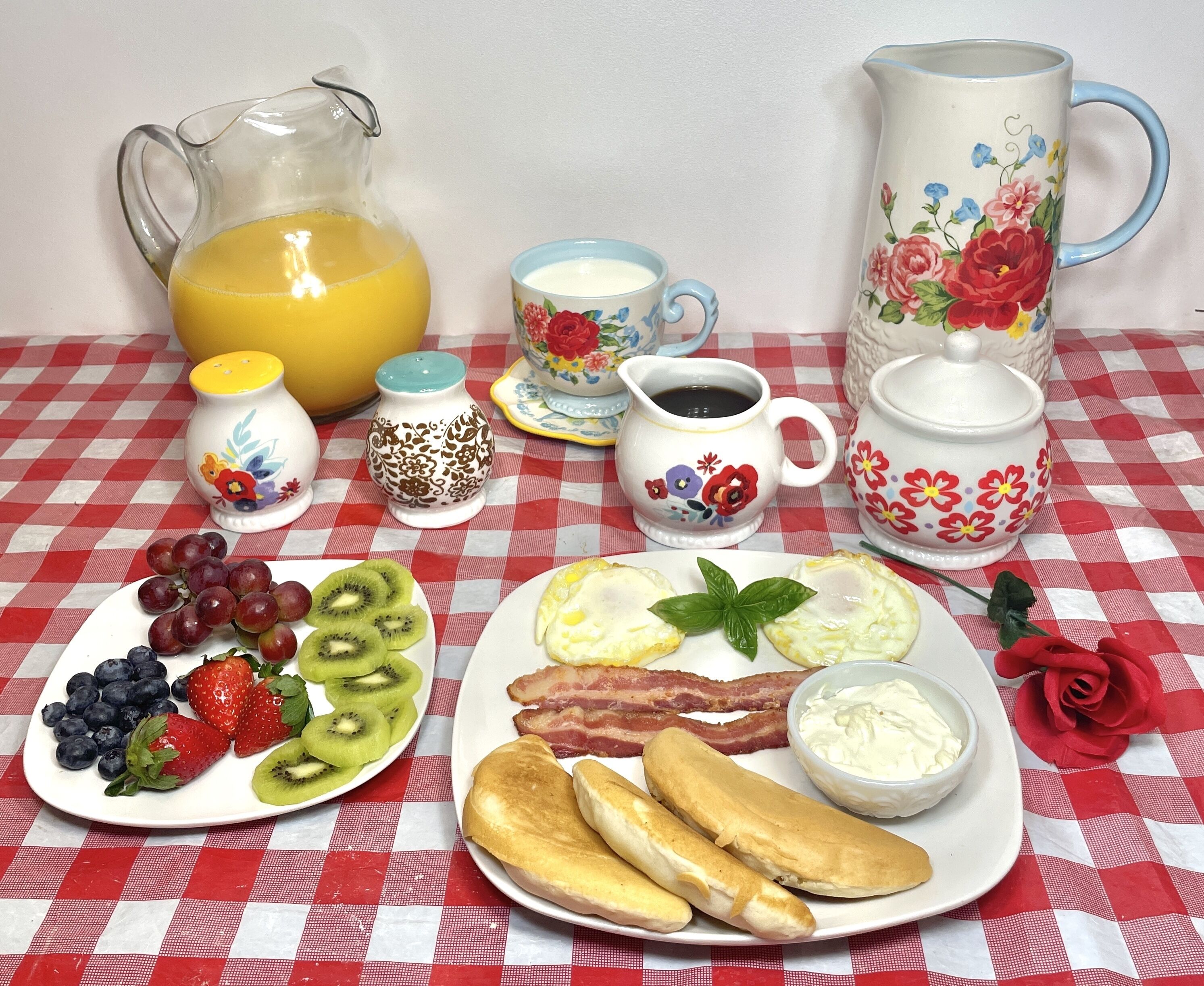 A breakfast table with a pitcher of orange juice, a cup of coffee, a teapot, a sugar bowl, a creamer, a plate of eggs, bacon, toast, and a plate of fruit.