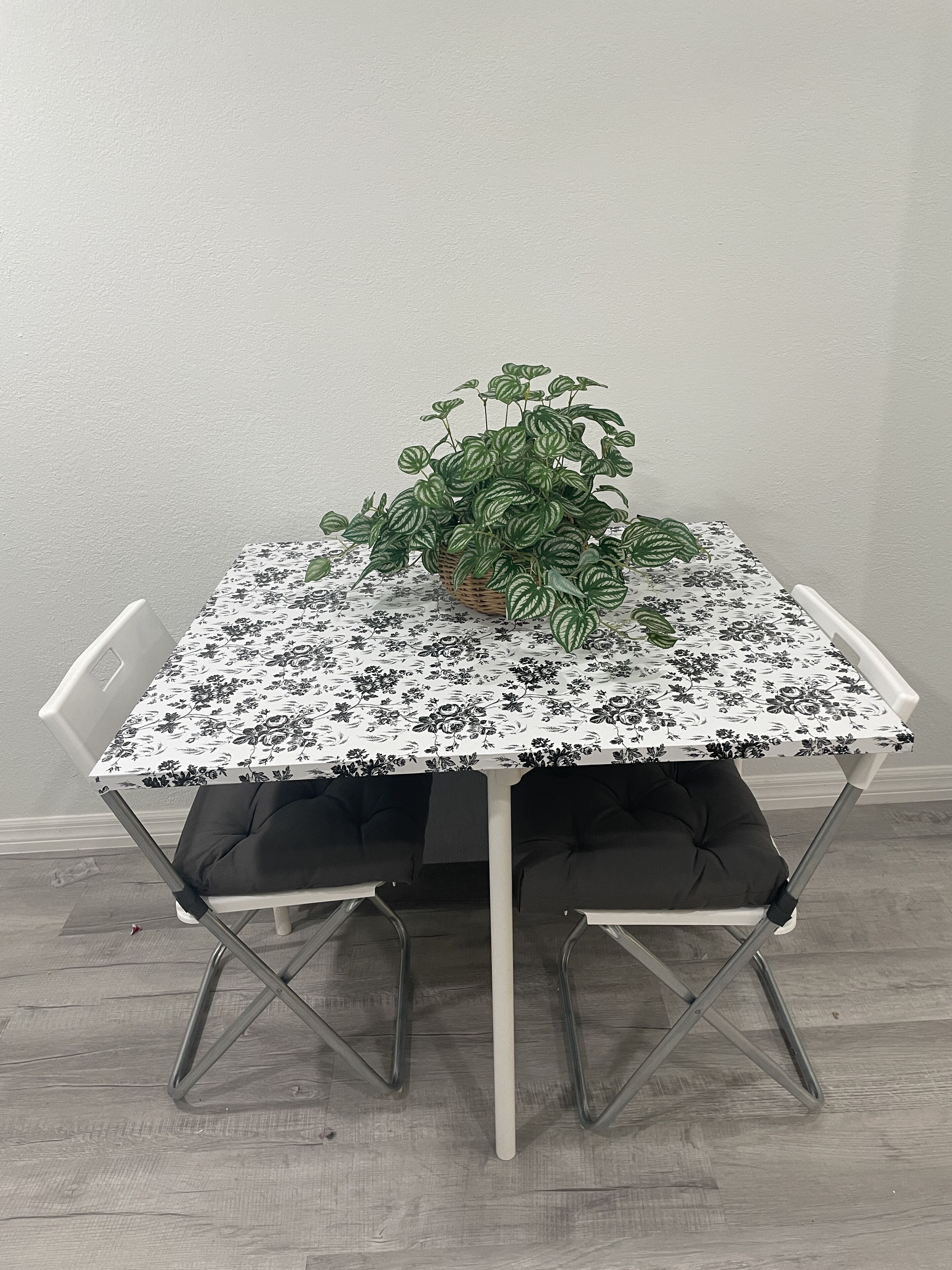 Une table à manger recouverte d’une nappe à motifs floraux, entourée de chaises blanches avec des coussins noirs.