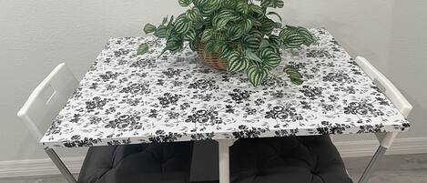 Une table à manger recouverte d’une nappe à motifs floraux, entourée de chaises blanches avec des coussins noirs.