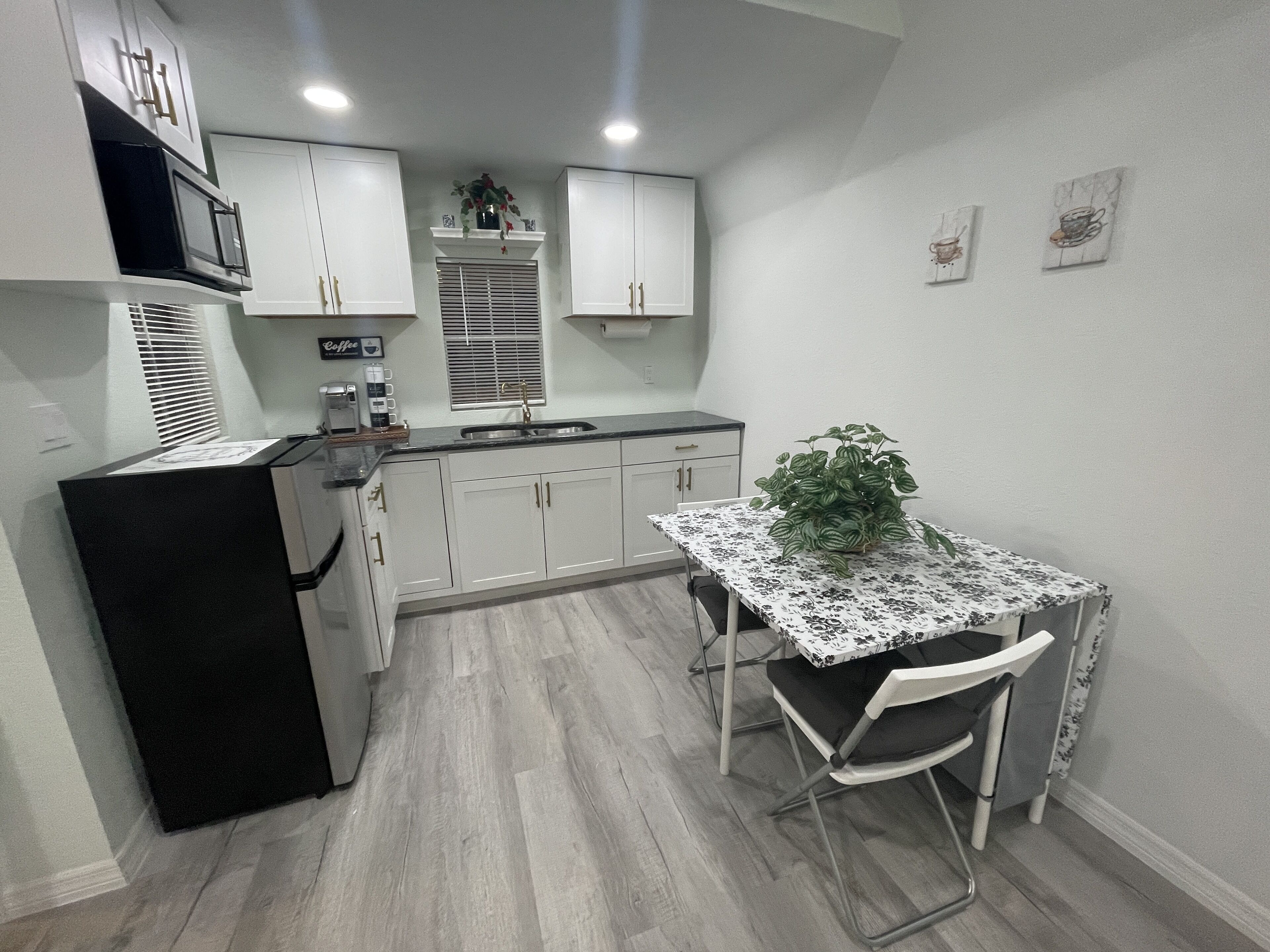Une cuisine compacte avec des meubles blancs, un réfrigérateur noir, une machine à café et une table à manger recouverte d’une nappe à motifs floraux.