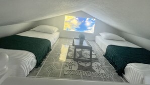 Ein kleines, gemütliches Schlafzimmer im Dachgeschoss mit zwei Betten, einem kleinen Tisch und einem Fenster mit Blick zum Himmel.