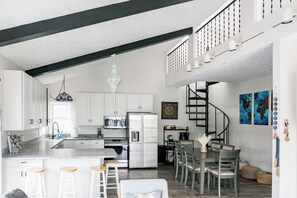 A modern kitchen with white cabinets, a central island with stools, a stainless steel refrigerator, and a dining area with a spiral staircase. - Peaceful Lakefront Getaway with Ping Pong on Apple Valley Lake (Howard)