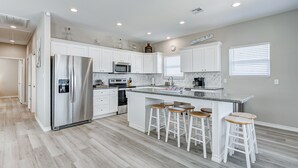 A modern kitchen with white cabinetry, a stainless steel refrigerator, a microwave, and a large island with bar stools.