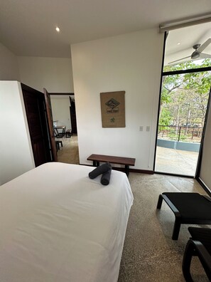 A hotel room with a large bed, a bench, a desk, and a view of the outdoors through a glass door. - Modern Beach Home with Pool & Rooftop - 150 metros (Santa Cruz, Playa Avellanas)