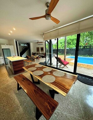 A modern kitchen with a wooden table and bench, large windows, and a view of a swimming pool. - Modern Beach Home with Pool & Rooftop - 150 metros (Santa Cruz, Playa Avellanas)