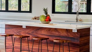 A modern kitchen with a marble island, wooden stools, and a fruit bowl. - Modern Beach Home with Pool & Rooftop - 150 metros (Santa Cruz, Playa Avellanas)