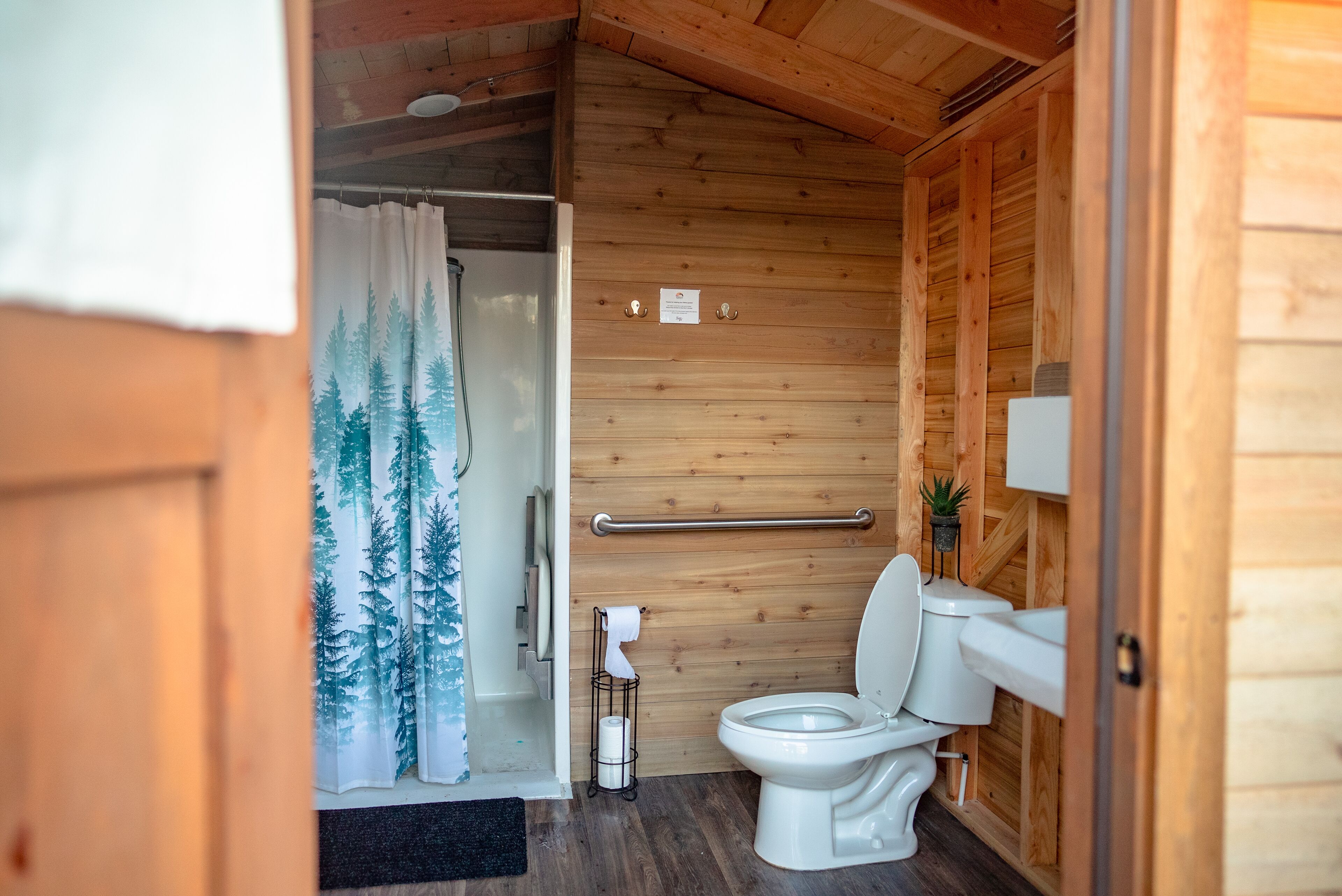 A compact wooden bathroom with a toilet, a shower curtain with a forest design, and a towel rack.