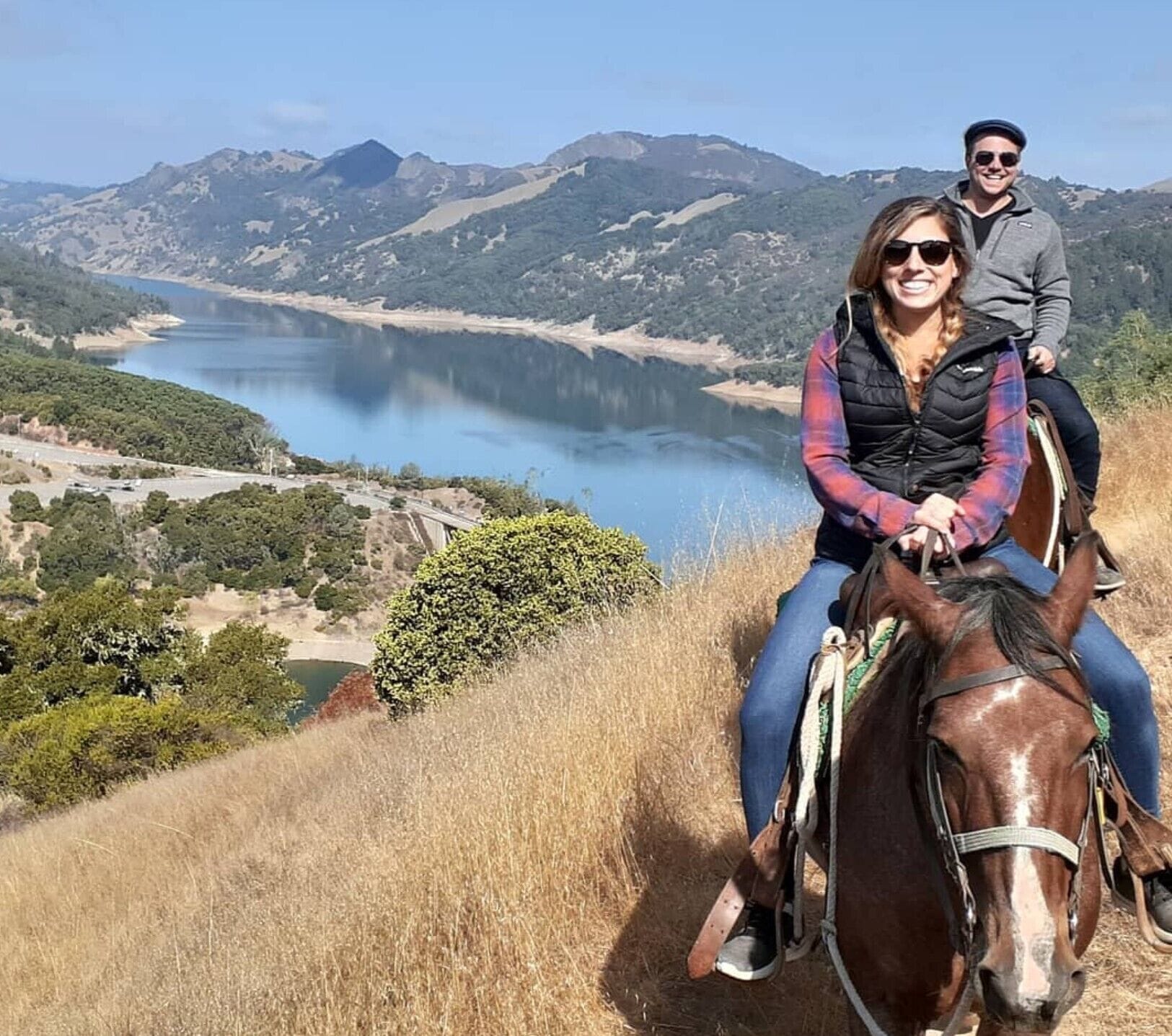 Duas pessoas cavalgam um cavalo numa colina, com um lago e montanhas ao fundo.