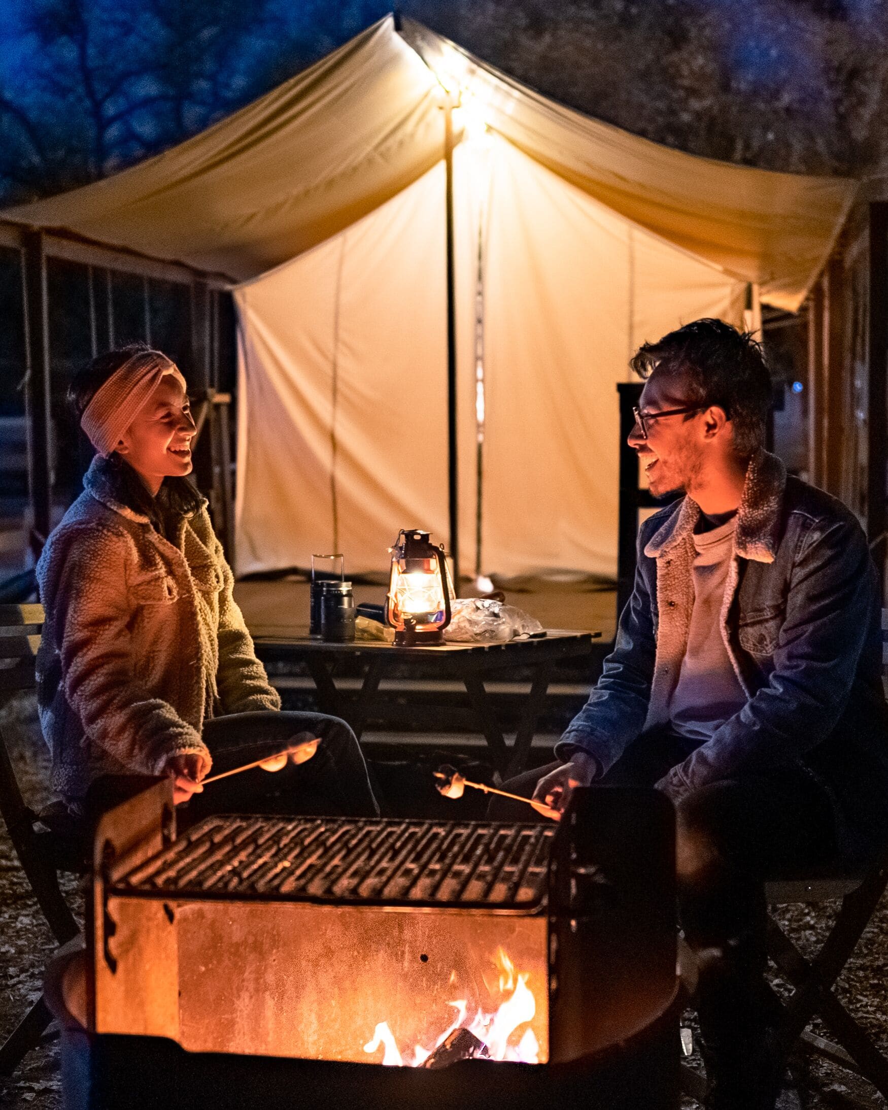 Twee mensen zitten bij een kampvuur, met een tent op de achtergrond.