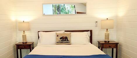 Une chambre avec un plafond en bois, un lit recouvert d’une couverture bleue, deux lampes de chevet, un miroir au-dessus du lit et trois paniers en osier placés sous le lit.