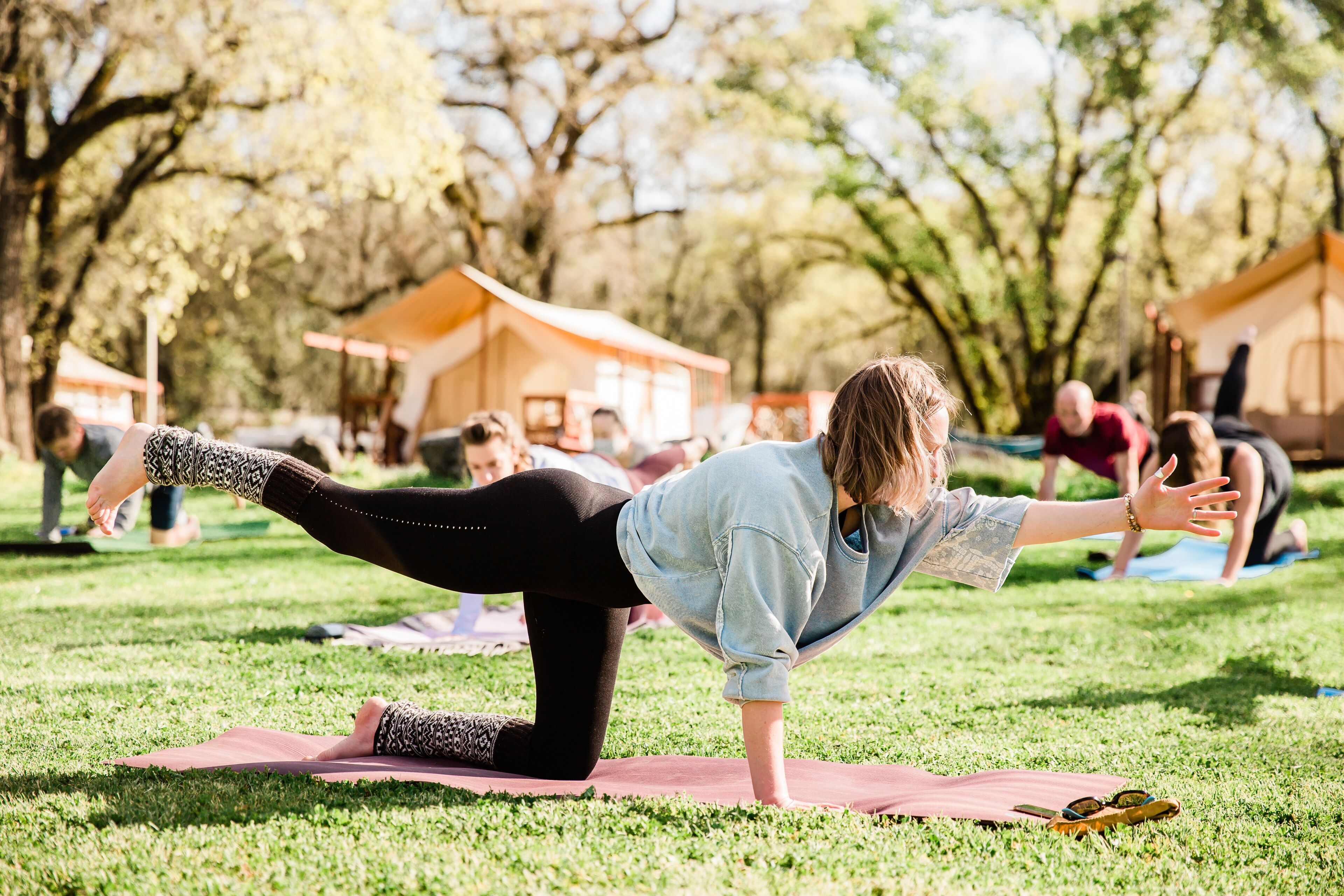 Iemand doet een yoga-houding op een mat in een park, met bomen en tenten op de achtergrond.