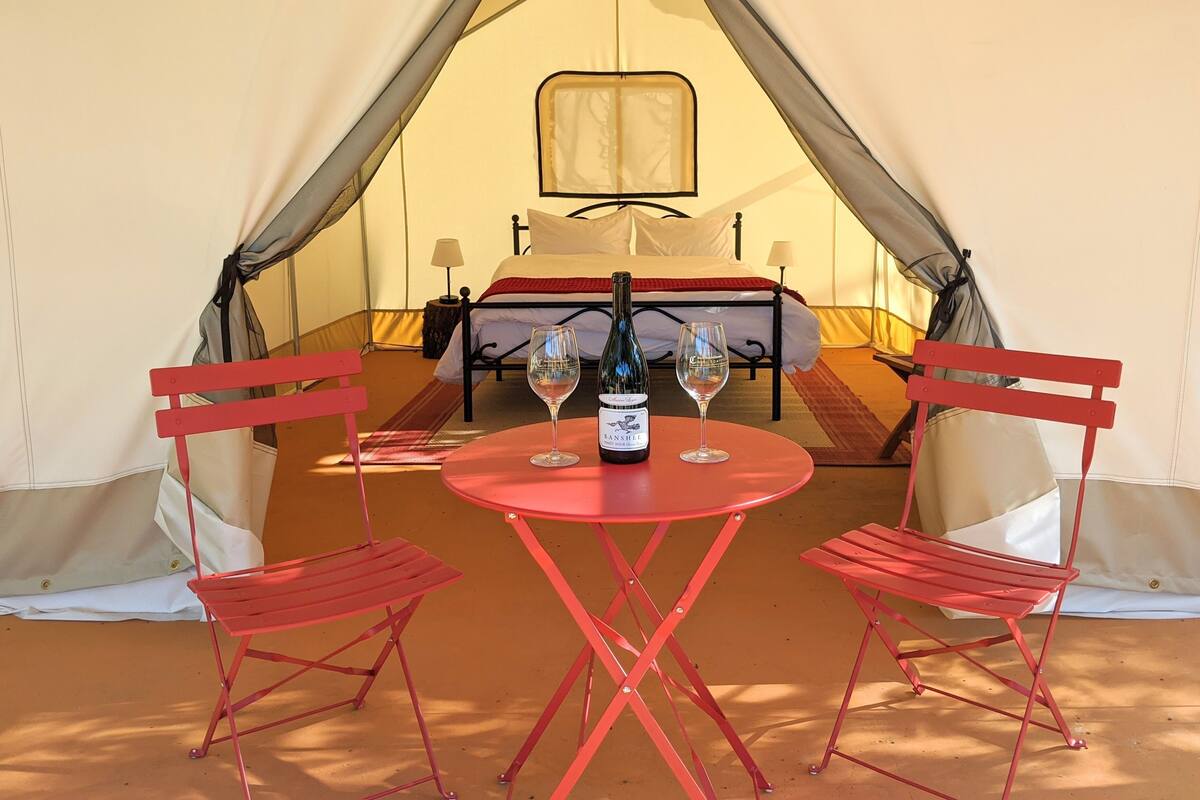 Een tent met een bed, twee rode stoelen, een tafel met een fles en glazen, en een lamp.