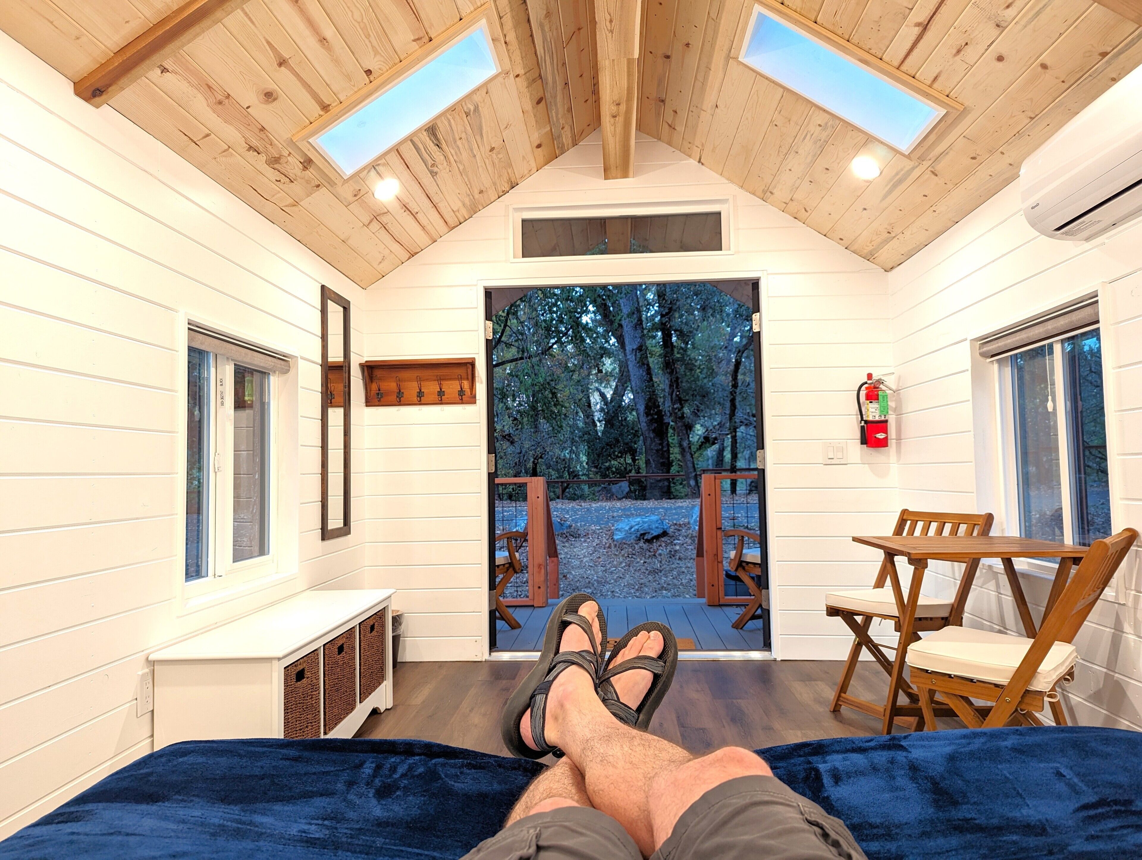 A cozy cabin interior with a bed, a table with chairs, a fire extinguisher, and a view of the forest through a door.