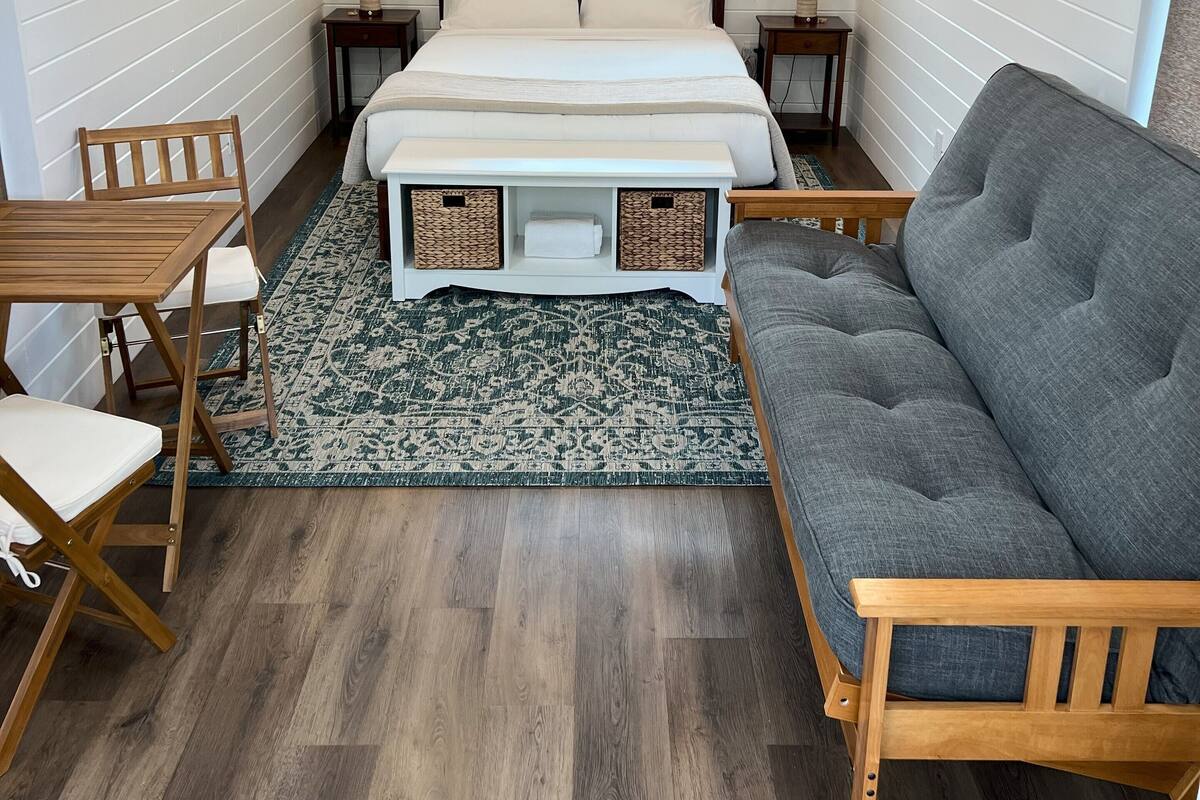 Een gezellig slaapkamer met een houten plafond, een bed met witte lakens, een grijze bank en een kleine tafel met stoelen.