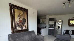 A living room with a grey sofa and ottoman, a painting on the wall, and a kitchen in the background. - VISTA AL MAR (Santa Marta)