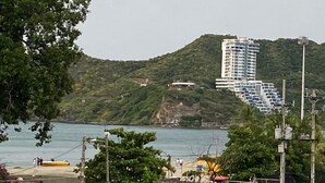A coastal area with a stone wall, boats, and a tall building on a hill. - VISTA AL MAR (Santa Marta)
