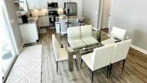 A modern kitchen with white cabinets, a central island, and a dining area with a glass table and white chairs. - Newburgh Newsie - 3BR, 2.5BA, Gym, Patio, New Construction in Amazing Location! (Newburgh)