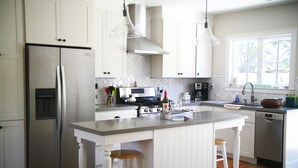 A modern kitchen with white cabinetry, stainless steel appliances, a central island with stools, and a window providing natural light. - Charming modern bungalow w/ hot tub steps away from the beach (Dillon Beach)